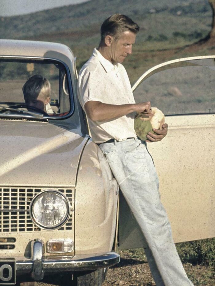 Man on vacation leaning against a vintage car peeling a coconut, capturing vacation pics from a time before cell phone cameras.