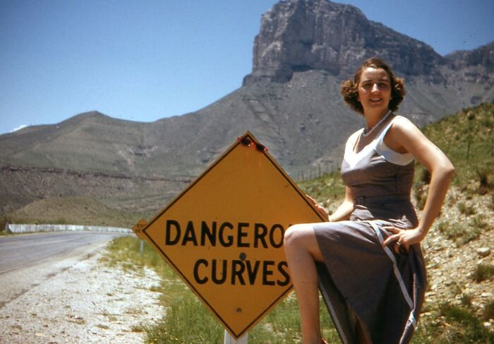 Vintage vacation pic of a woman sitting by a dangerous curves road sign in a mountainous desert landscape.