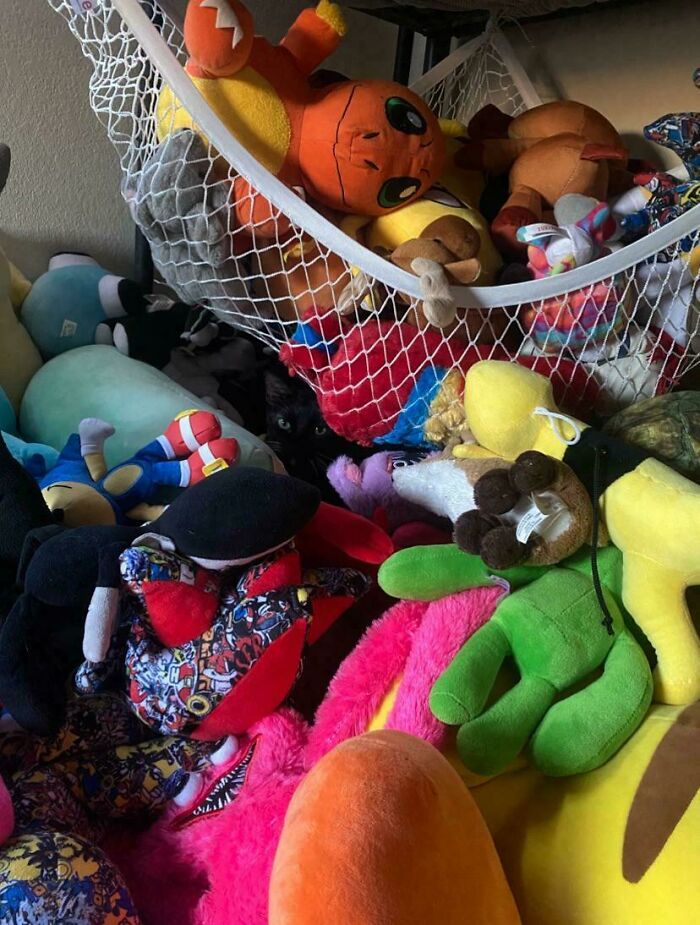 Black cat seamlessly merged into a pile of colorful stuffed toys blending perfectly with the background.