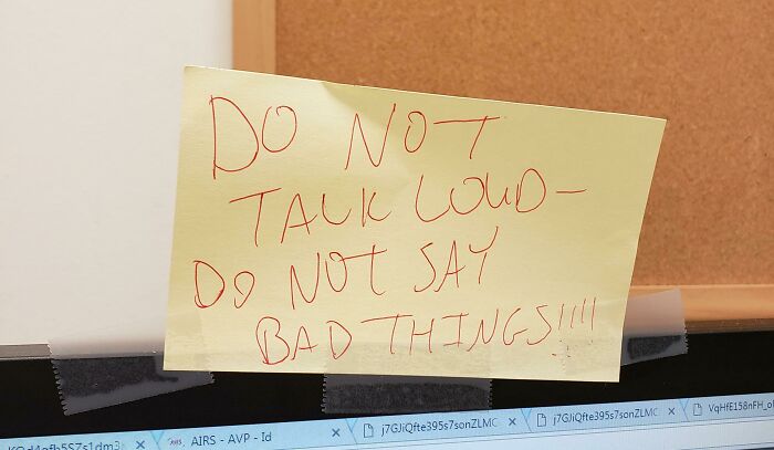 Yellow sticky note with a passive-aggressive message taped above computer screens in a coworker’s workspace.