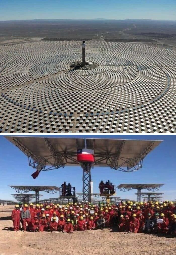 A massive solar power plant with workers in red uniforms and helmets showing human for scale perspective.