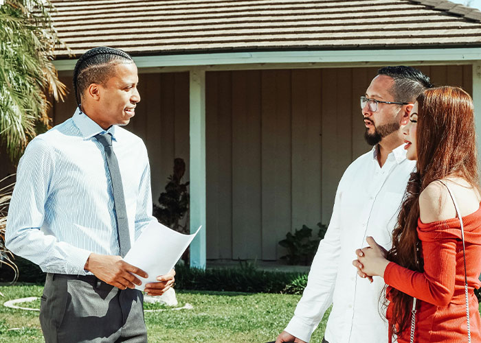 Real estate agent discussing property details outdoors with a couple near a suburban home and white oak trees.