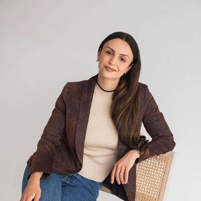Young woman with long dark hair wearing a brown blazer and beige top, seated on a chair in a relaxed pose.