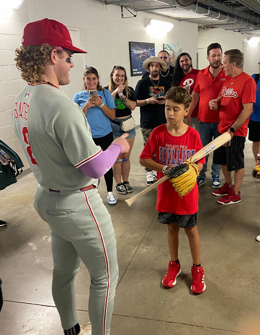 Baseball player signing autograph for young Phillies fan in hallway with crowd in background after game event.