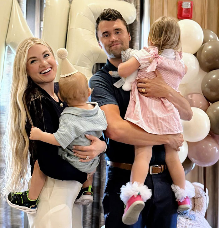 Charlie Kirk's wife smiling and holding a child, standing next to Charlie Kirk holding another child at a celebration.