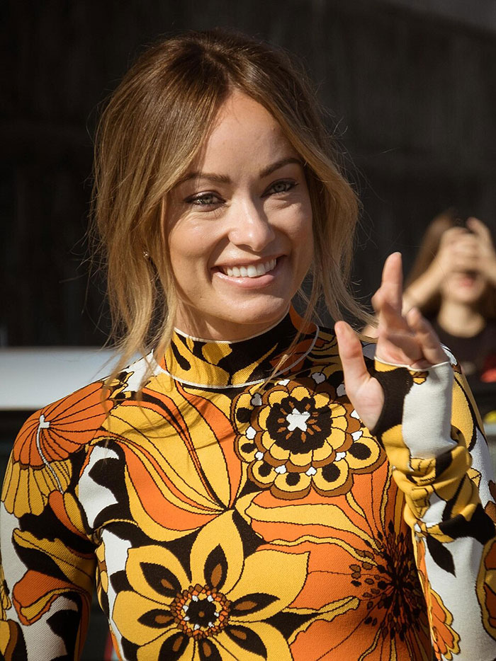 Olivia Wilde smiling and waving in a vibrant patterned outfit, showcasing her toned abs during New York Fashion Week.