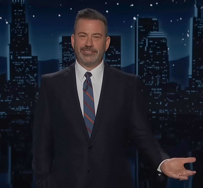 Jimmy Fallon speaking on stage in a suit and tie against a nighttime city skyline background during a show.