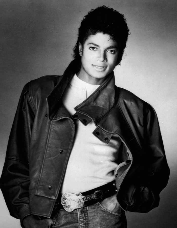 Michael Jackson in a black leather jacket and white shirt, posing confidently with hands in pockets in a classic black and white photo.