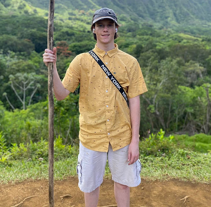 Young person standing outdoors in a yellow shirt holding a wooden stick amid lush greenery, related to Tyler Robinson's trans partner disappearance. Young person standing outdoors in a yellow shirt holding a wooden stick amid lush greenery, related to Tyler Robinson's trans partner disappearance.