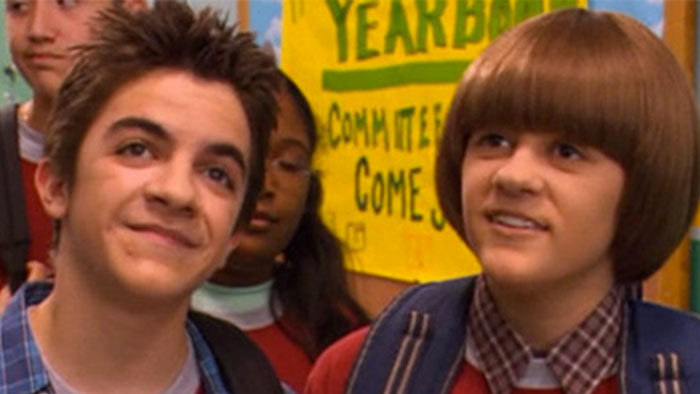 Two young actors from a Nickelodeon show smiling in a school hallway with a colorful yearbook sign in the background