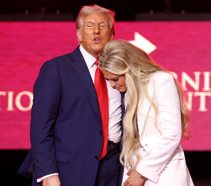 Elon Musk in a dark suit with red tie standing next to a woman with long blonde hair in a white outfit on stage.