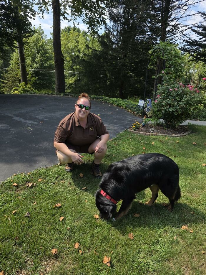 UPS driver smiling and posing outdoors with an adorable black dog on a grassy lawn near a driveway.