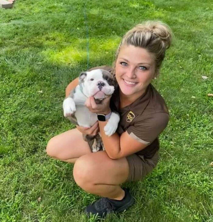 UPS driver in uniform smiling and holding an adorable bulldog puppy while crouching on green grass outdoors