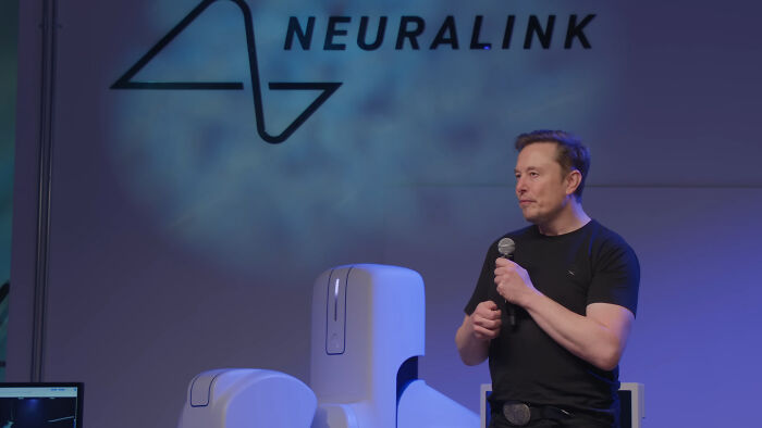 Elon Musk speaking on stage with Neuralink branding behind him, holding a microphone during a presentation.