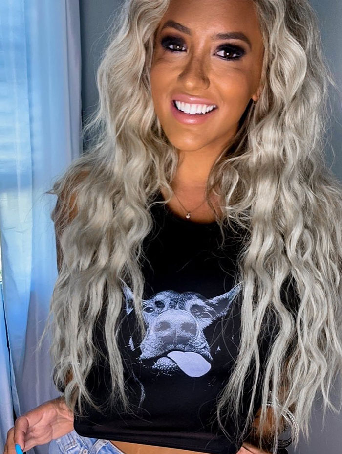 Blonde model with long wavy hair smiling, wearing a black shirt, involved in big breasts discrimination claim.