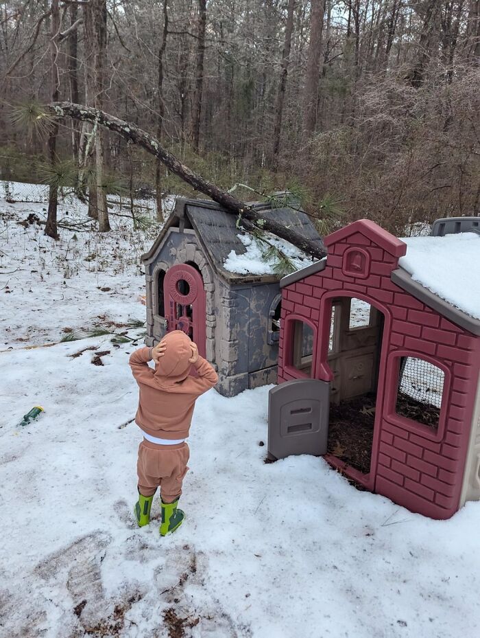 Child in rain boots looking at a fallen tree on a playhouse roof, capturing wholesome moments in a snowy backyard setting.