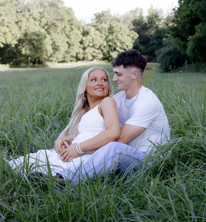 Cheerleader appearing all glammed up in court, sitting outdoors with partner in a grassy field, smiling and embracing.