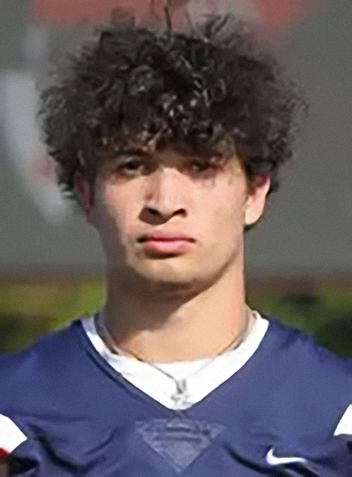 Young man with curly hair in a blue sports jersey, posing outdoors with a serious expression and blurred background.