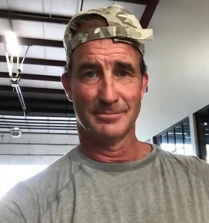 Middle-aged man wearing a camo cap and gray shirt indoors, related to husband of woman caught with CEO amid divorce news. Middle-aged man wearing a camo cap and gray shirt indoors, related to husband of woman caught with CEO amid divorce news.
