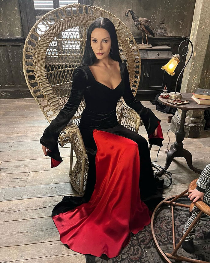 Catherine Zeta-Jones sitting in a wicker chair wearing a black and red dress in a rustic room setting.