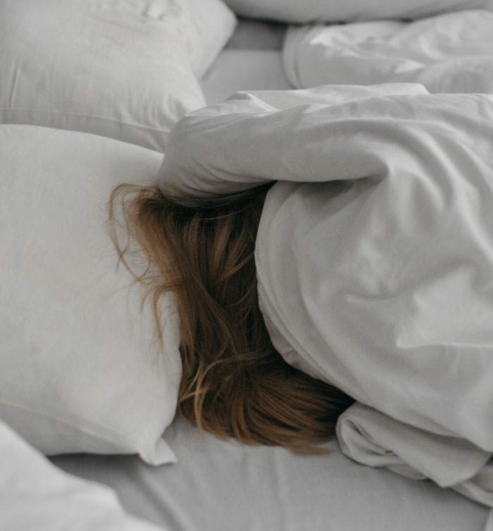 Person hiding under white blankets in bed, evoking creepy moments experienced while home alone.