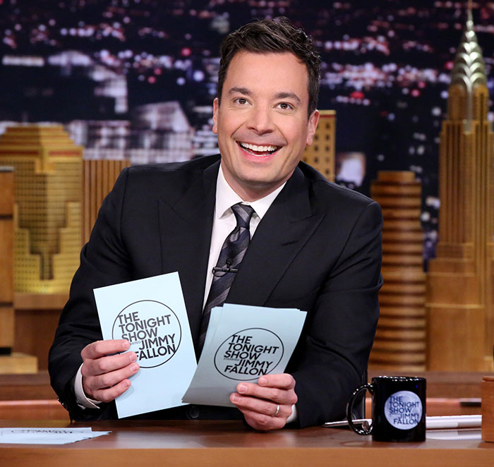 Jimmy Fallon smiling on The Tonight Show set, holding cards and wearing a black suit with cityscape background.