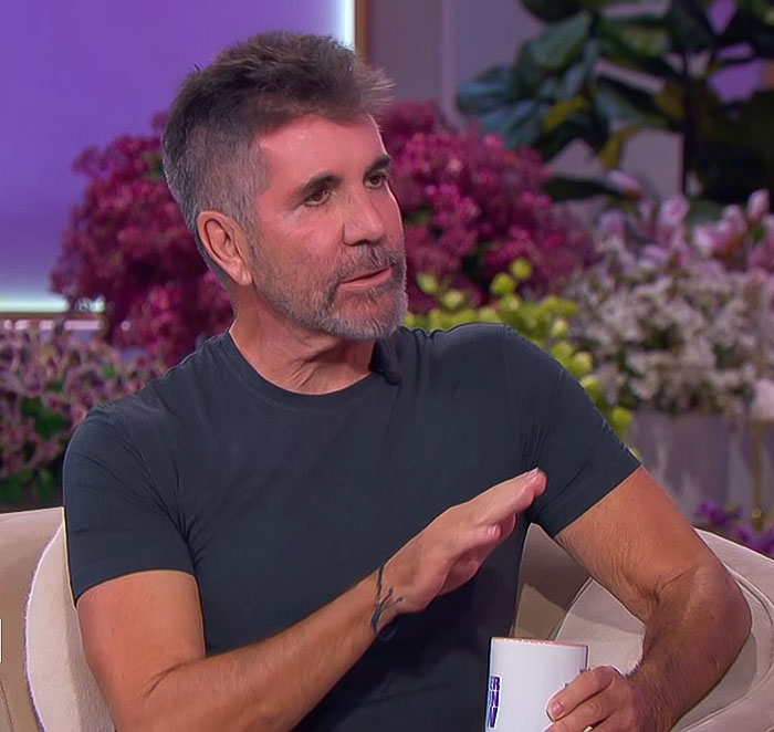 Simon Cowell speaking during an interview with flowers in the background after a bike accident recovery.