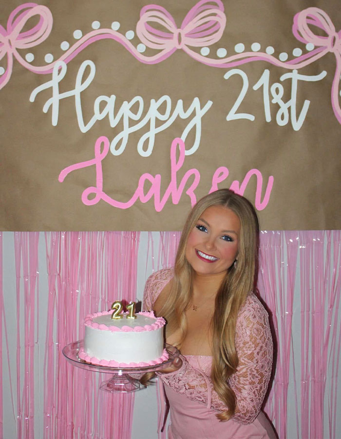 Young cheerleader smiling and holding a pink and white birthday cake celebrating her 21st birthday indoors.