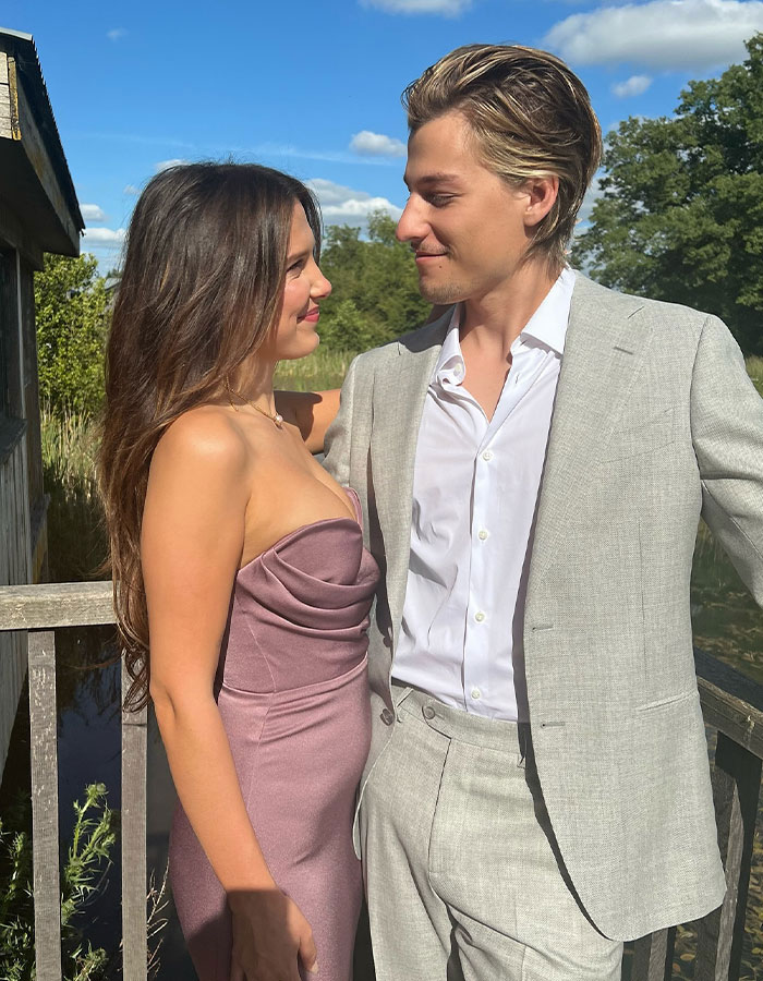 Couple standing outdoors on a wooden bridge, dressed elegantly in a pink dress and light gray suit under a blue sky.