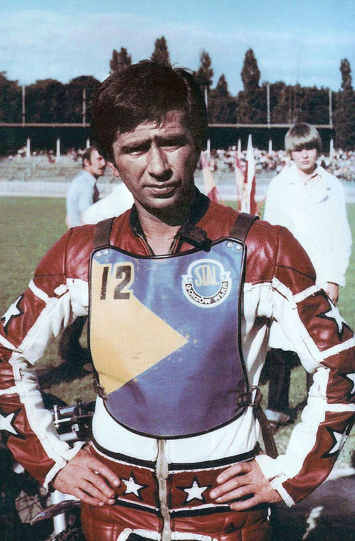 Male celebrity wearing vintage racing gear with number 12, standing on a track with a crowd and trees in the background.