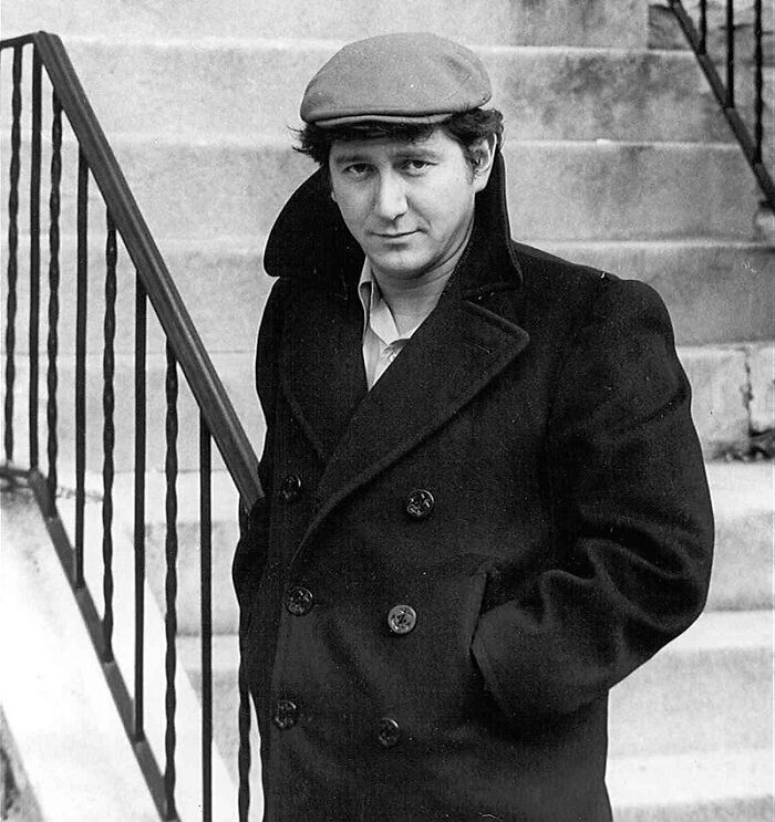 Black and white photo of a man in a coat and cap standing by stairs, representing American folk heroes in history.