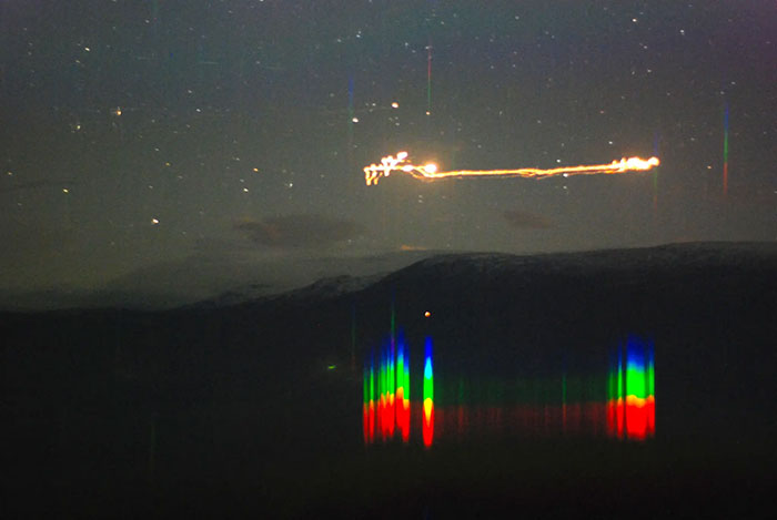 Night sky showing stars and a mysterious glowing light trail with colorful reflections over a dark landscape.