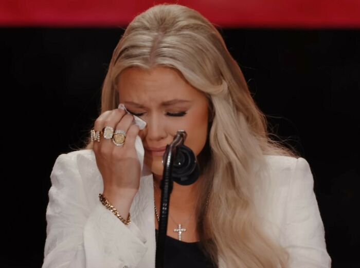 Woman wiping tears during heated debate about Erika Kirk and feminist movement at public event.