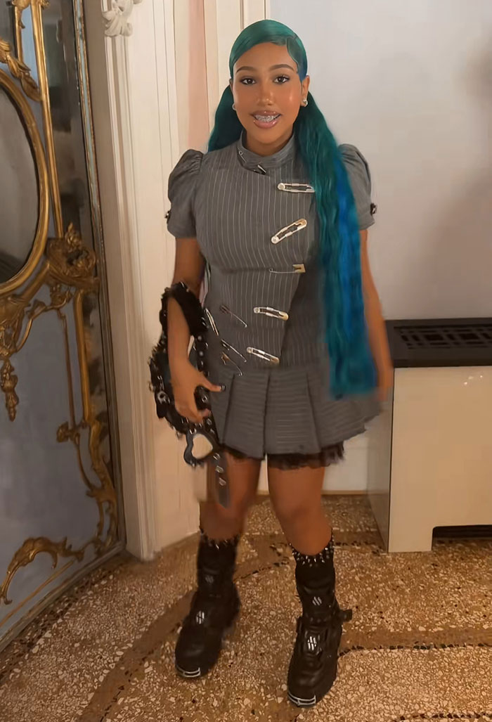 Young girl with long blue hair wearing a gray pinstripe dress and black boots, standing indoors in a decorative room.