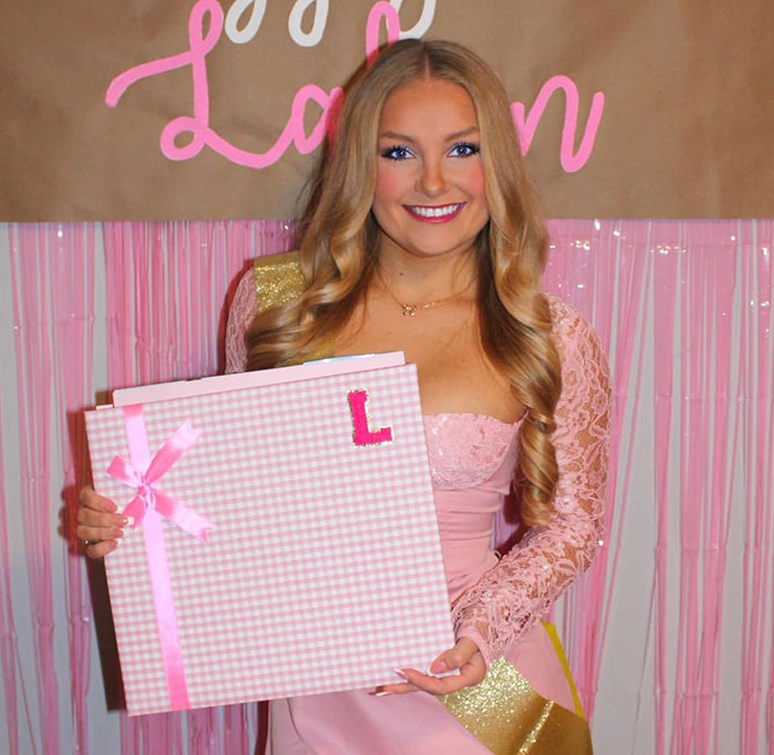 Cheerleader in pink dress smiling and holding a large pink gift box with ribbons at an event background.