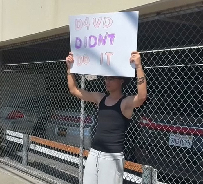 Young man holding a sign near an abandoned Tesla linked to singer, amid decomposing body discovery controversy.