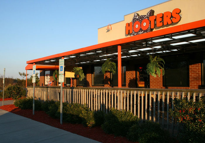 Fachada de restaurante Hooters al atardecer, con plantas colgantes y señales de estacionamiento para discapacitados.
