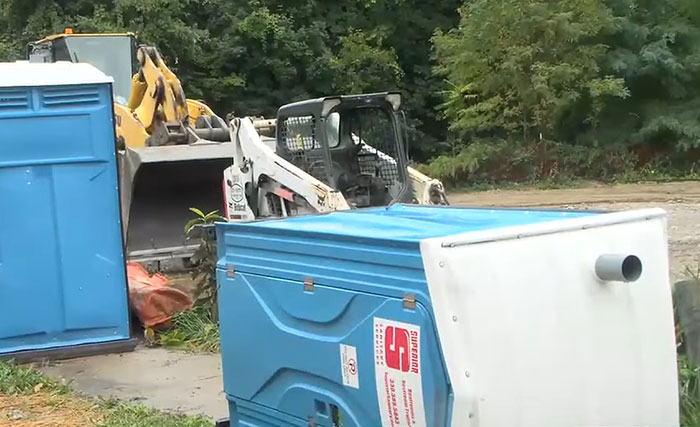 Blue porta-potty tipped over near construction equipment in an outdoor area, related to cop shooting teens over porta-potty prank.