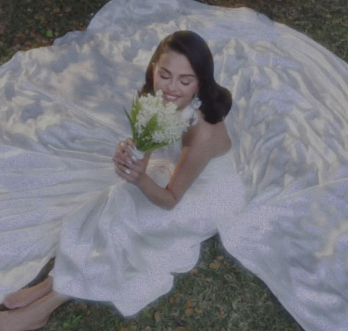 Bride smiling while holding flowers, sitting on grass with a flowing wedding dress at a celebrity wedding event.