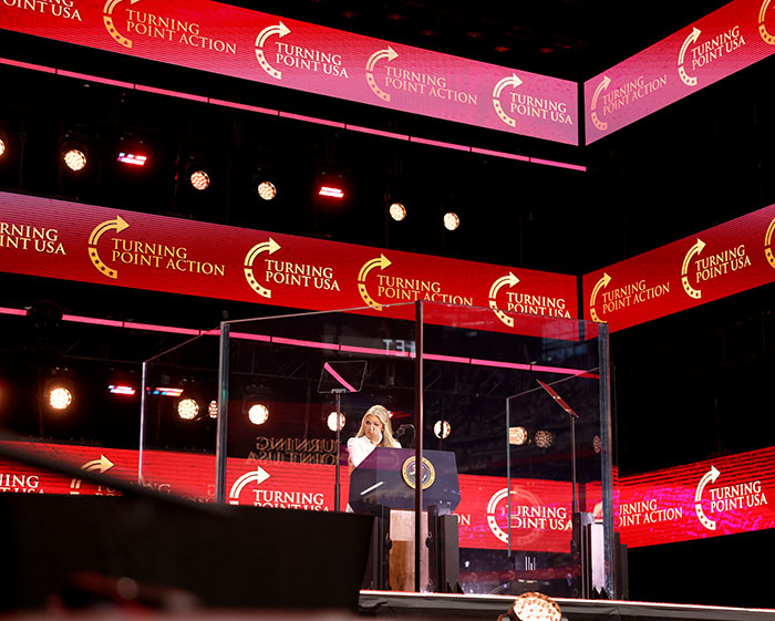 Speaker at Turning Point USA event surrounded by red and pink digital signs, related to Elon Musk accused of hypocrisy.