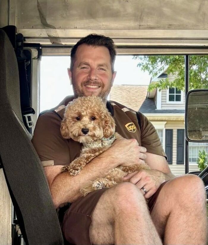 UPS driver smiling inside delivery truck, holding an adorable curly-haired pet dog in his arms.