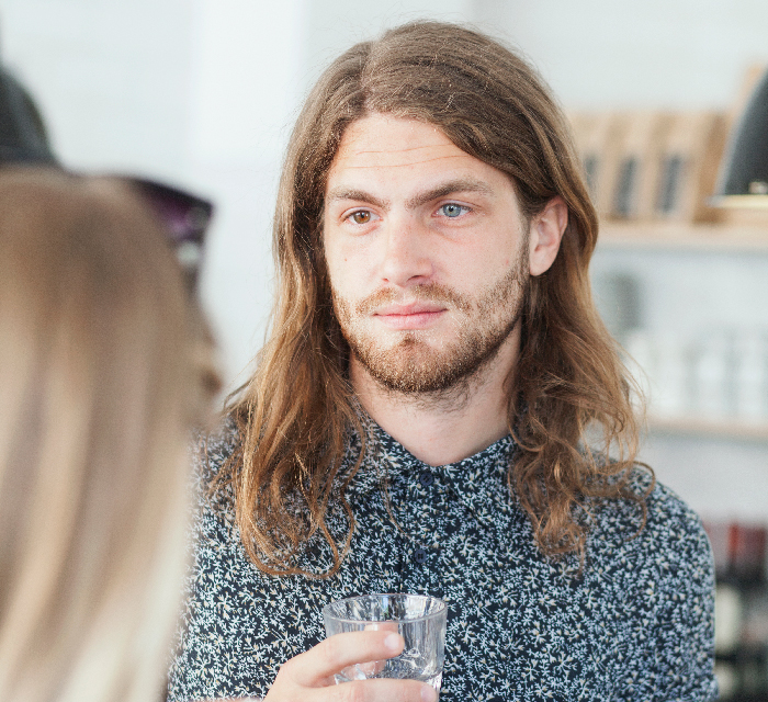 Young man with long hair and beard holding glass of water, facing a blurred person in an indoor setting.