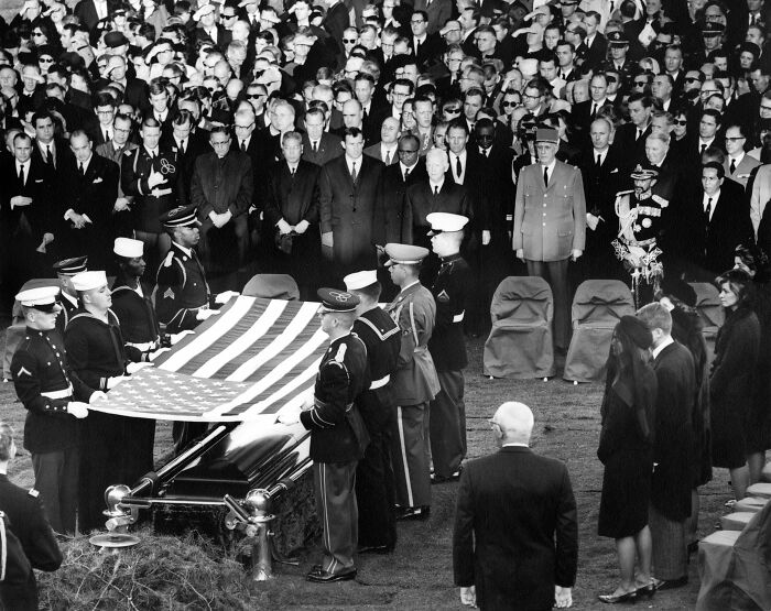 Military honor guard folding American flag at a large funeral with mass fury compared to Martin Luther King and John F. Kennedy.