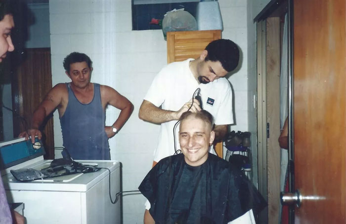 Man diagnosed with testicular cancer gets haircut while friends watch in a small room with vintage appliances.