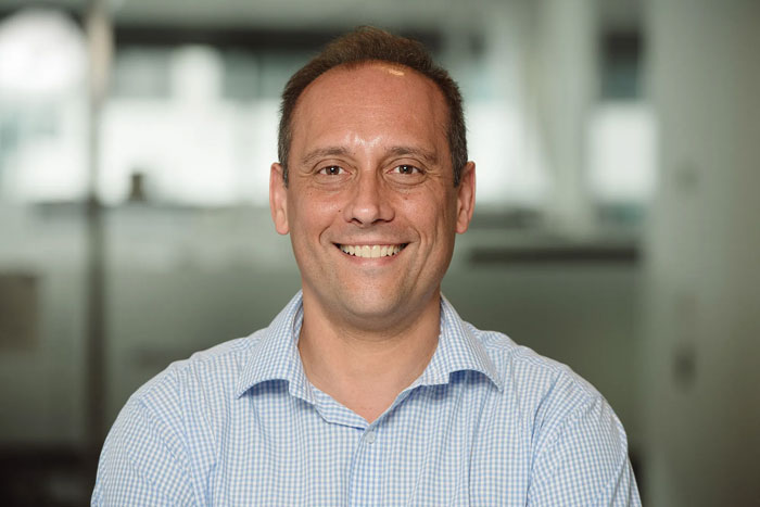 Man diagnosed with testicular cancer smiling in a light blue checked shirt against a blurred background.