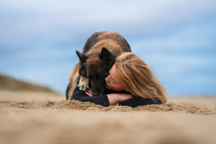 Woman lying on sand embracing her dog in a heartwarming pet photo from the 2025 International Pet Photography Awards.