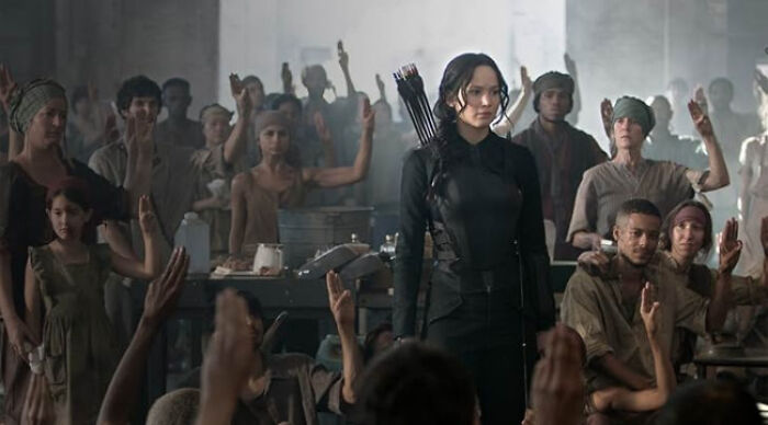 A female archer in black stands while a crowd of people raises their hands in a beloved movie tropes scene.
