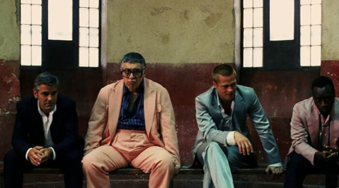 Four men dressed in suits sitting on a bench in a dimly lit room, representing beloved movie tropes.