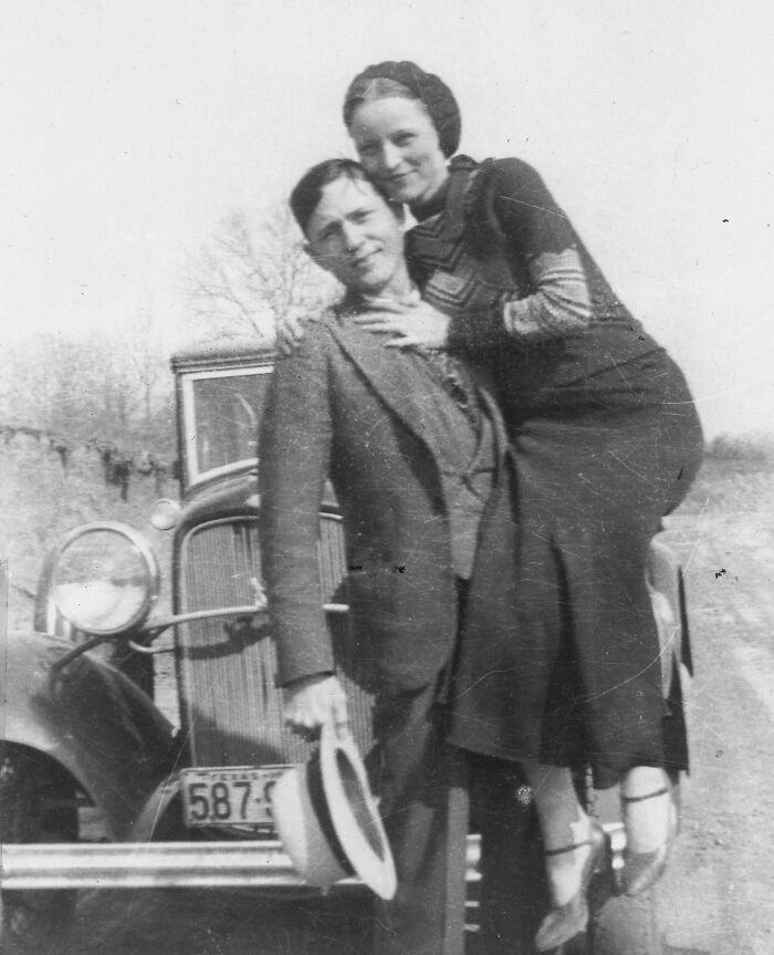 Vintage black and white photo of an American folk hero couple posing by an old car in a rural setting.