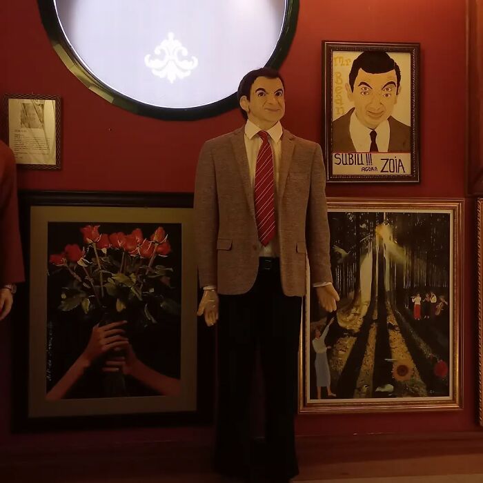 Wax sculpture of a famous figure in a suit and tie, surrounded by framed paintings, showcasing creepy and hilarious artistry.